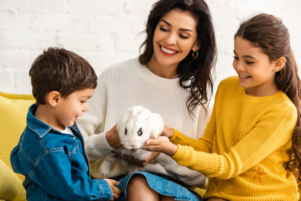 счастливые дети и веселая мать сидит на диване с белым смешным кроликом
 - Фото, изображение
