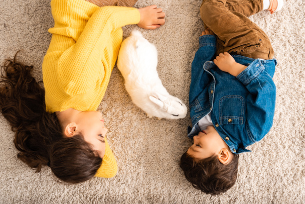 top view of cute brother and sister lying on floor near white funny rabbit - Foto, Imagen