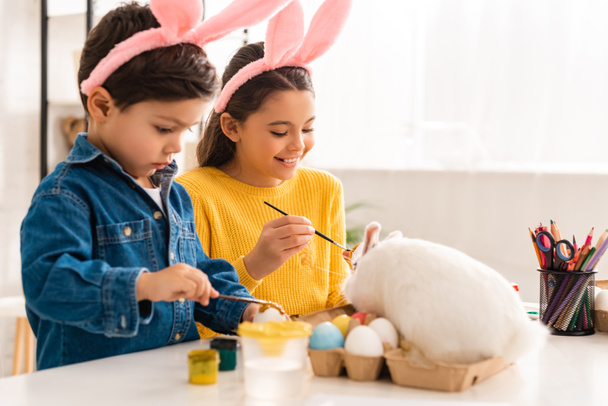 imádnivaló testvér és nővér nyuszifülekben húsvéti tojásokat festenek, miközben az asztalnál ülnek a fehér nyúl közelében - Fotó, kép