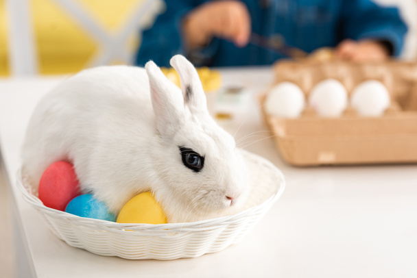 selektywne skupienie białego królika w wiklinach na stole obok chłopca malującego pisanki - Zdjęcie, obraz