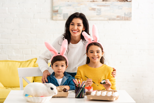 gelukkig vrouw en kinderen in konijnenoren glimlachen op camera terwijl schilderen paaseieren in de buurt van wit konijn in wicker - Foto, afbeelding