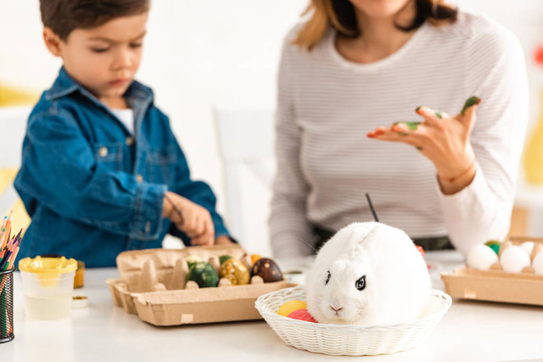 selective focus of adorable boy painting Easter eggs while sitting near mother and white rabbit in wicker - Fotoğraf, Görsel