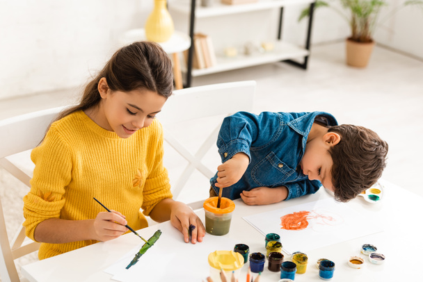 lindo chico mezcla pinturas en contenedor mientras está sentado cerca de hermana dibujo con pincel
 - Foto, imagen