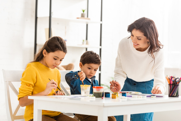 happy woman standing near table and drawing with paints together with children - Photo, Image