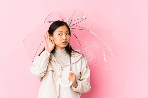 Young chinese woman holding a umbrella isolated trying to listening a gossip. - Fotoğraf, Görsel