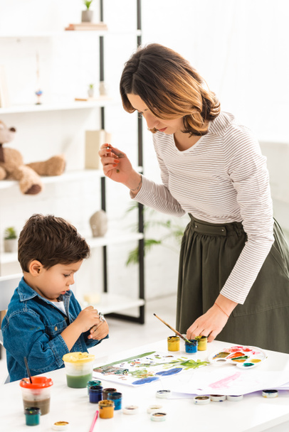 young mother standing near table while cute son holding container with paints - Photo, Image