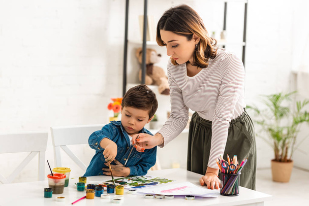 young mother holding paintbrush while son drawing with paints - Foto, afbeelding