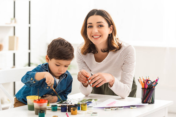 glückliche Frau blickt in die Kamera, während sie mit ihrem Sohn mit Farben zeichnet - Foto, Bild