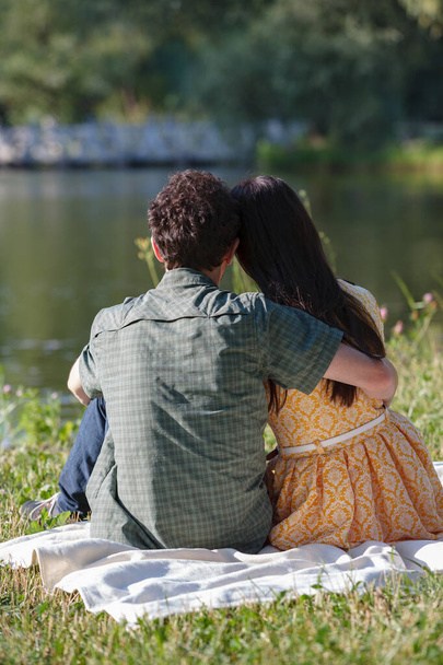 On white plaid sit back young couple. They look at lake. A man embraces a woman. - Photo, Image