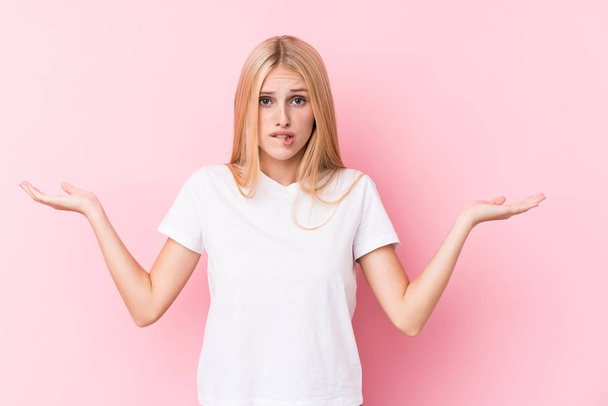 Young blonde woman on pink background confused and doubtful shrugging shoulders to hold a copy space. - Foto, imagen