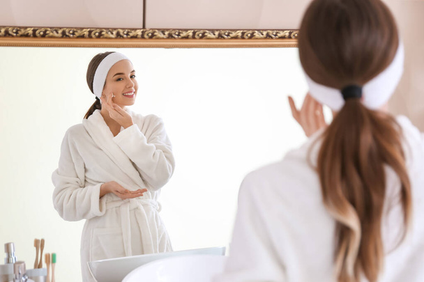 Beautiful young woman applying cream on her face near mirror in bathroom - Фото, изображение