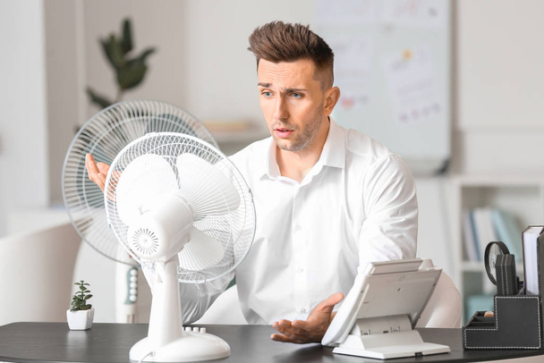 Jeune homme souffrant de chaleur près d'un ventilateur électrique au bureau
 - Photo, image