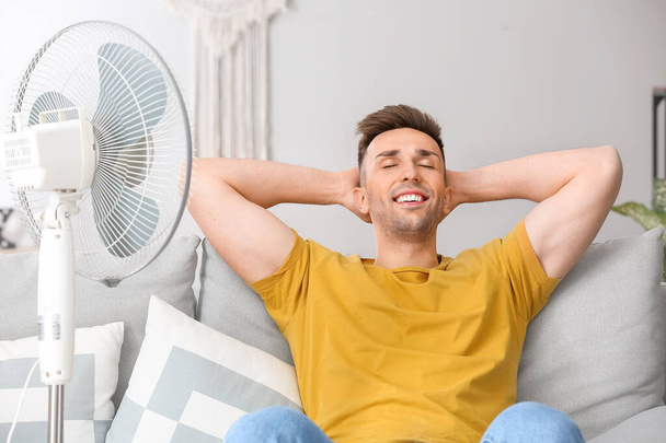 Hombre joven usando ventilador eléctrico durante la ola de calor en casa
 - Foto, Imagen