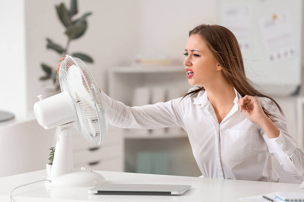 Розлючена жінка, яка страждає від спеки біля електричного вентилятора в офісі
 - Фото, зображення