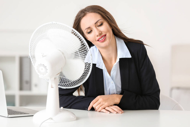Молода жінка використовує електричний вентилятор під час теплової хвилі в офісі
 - Фото, зображення