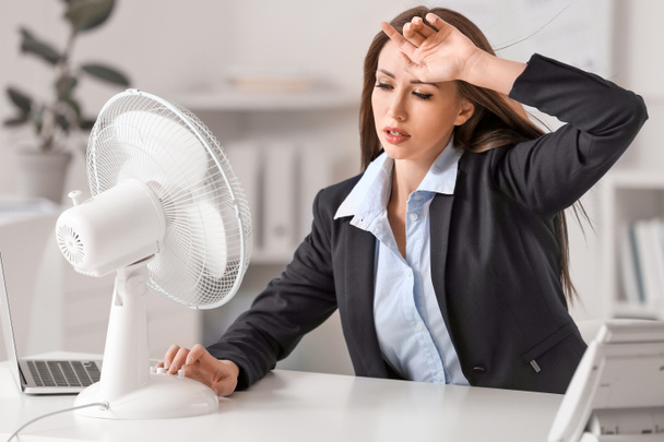 Young woman using electric fan during heatwave in office - 写真・画像