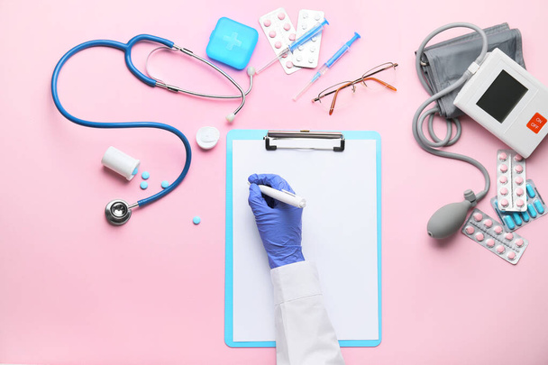 Χέρι του γιατρού που γράφει στο πρόχειρο, σφυγμομανόμετρο και χάπια στο φόντο χρώμα - Φωτογραφία, εικόνα
