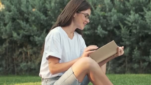 Beautiful young girl wearing glasses is reading a book while sitting outside in the city park in the summer - Séquence, vidéo