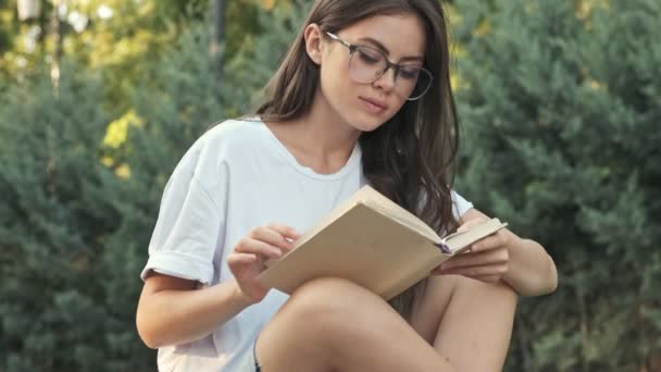 Charming young girl wearing glasses is reading a book then look to the camera with a smile outside in the city park in the summer - Záběry, video