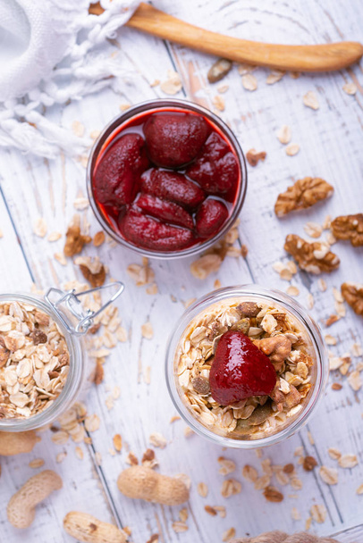 Potje zelfgemaakte granola met yoghurt en ingeblikte aardbeien op witte houten achtergrond bovenaanzicht - Foto, afbeelding