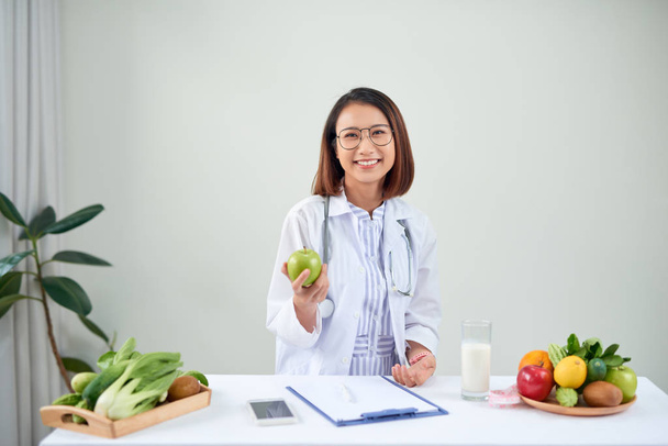 Usmívající se odbornice na výživu ve své kanceláři ukazuje zdravou zeleninu a ovoce, zdravotní a dietní koncepci. - Fotografie, Obrázek