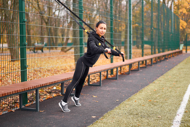 Girl athlete functional training on sportground. Mixed race young adult woman do workout with suspension system. Healthy lifestyle. Stretching outdoors playground. - 写真・画像