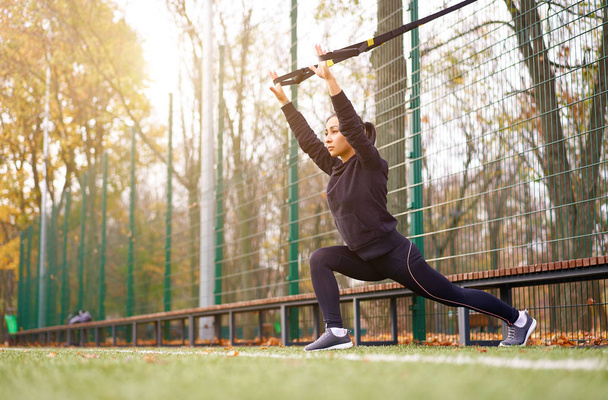 Girl athlete functional training on sportground. Mixed race young adult woman do workout with suspension system. Healthy lifestyle. Stretching outdoors playground. - Foto, imagen