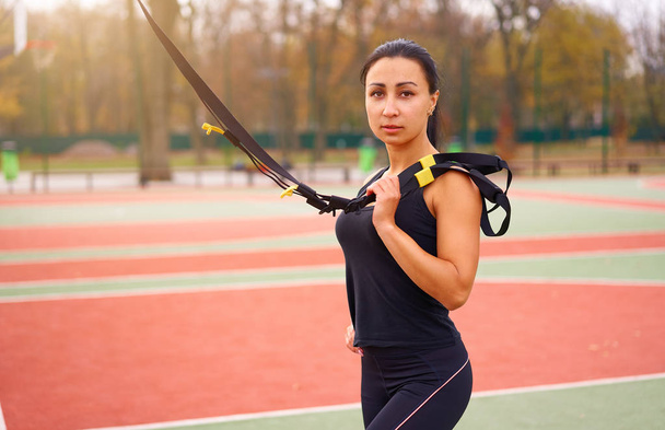 Girl athlete functional training on sportground. Mixed race young adult woman do workout with suspension system. Healthy lifestyle. Stretching outdoors playground. - Foto, afbeelding