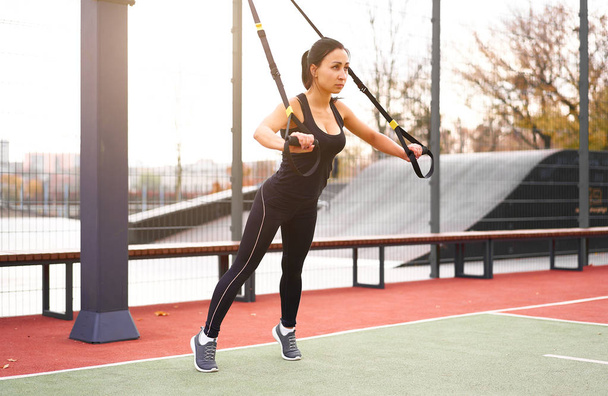 Girl athlete functional training on sportground. Mixed race young adult woman do workout with suspension system. Healthy lifestyle. Stretching outdoors playground. - Foto, Bild