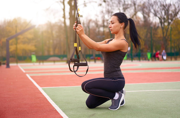 Girl athlete functional training on sportground. Mixed race young adult woman do workout with suspension system. Healthy lifestyle. Stretching outdoors playground. - Photo, image