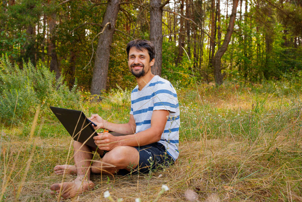 Młody brunetka z brodą siedzi na trawie z laptopem. Student pracuje na zewnątrz - Zdjęcie, obraz