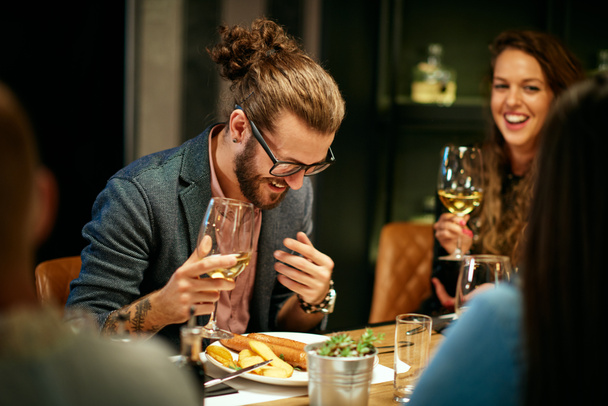 Νεαρός όμορφος καυκάσιος χίπστερ κάθεται με φίλους στο εστιατόριο, κρατώντας ένα ποτήρι κρασί και γελώντας. - Φωτογραφία, εικόνα