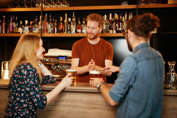 Fröhliche Freunde lehnen an der Theke, trinken Bier und unterhalten sich mit dem Barkeeper. Ausgehen. - Foto, Bild
