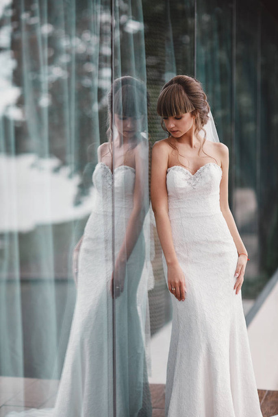 Την ημέρα του γάμου. Πορτραίτο νύφης σε λευκό φόρεμα έξω από το παράθυρο. Ομορφιά πορτρέτο της νύφης φορώντας νυφικό μόδας με πολυτέλεια απόλαυση μακιγιάζ και χτένισμα. - Φωτογραφία, εικόνα