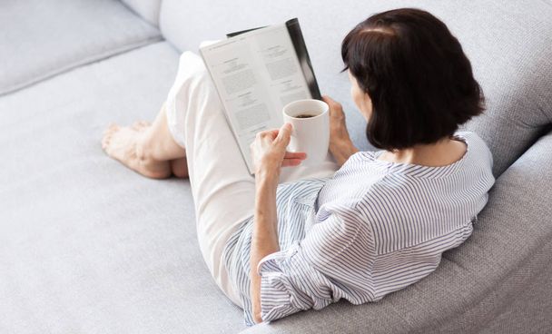 brunetka w średnim wieku kobieta w okularach na szarej kanapie czytania - Zdjęcie, obraz