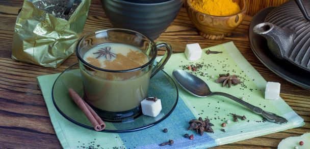 Τσάι Μασάλα. Ένα φλιτζάνι παραδοσιακό ινδικό τσάι masala τσάι με συστατικά για να προετοιμάσει. Κανέλα, γλυκάνισο, ζάχαρη, μαύρο τσάι, πιπέρι, γαρύφαλλο, κουρκούμη σε ξύλινο τραπέζι. - Φωτογραφία, εικόνα