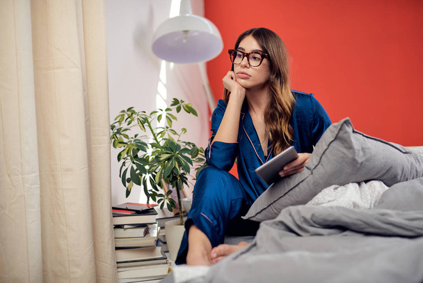 Bored cute caucasian brunette in pajamas with eyeglasses sitting on bed and holding tablet in the morning. - Foto, Imagem
