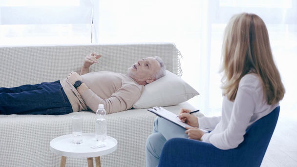 mature man lying on couch and talking to psychologist writing on clipboard - Footage, Video