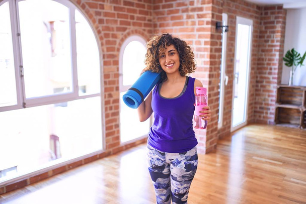 junge schöne arabische Sportlerin lächelt glücklich. stehend mit Sportmatte und Wasserflasche vor dem Yoga-Kurs im Fitnessstudio - Foto, Bild