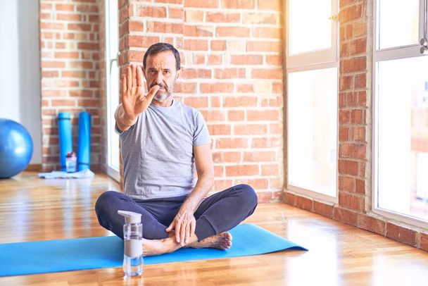 gutaussehender Sportler mittleren Alters, der auf einer Matte sitzt und im Fitnessstudio Stretching Yoga-Übungen macht, wobei er mit der Handfläche aufhört zu singen. Warnausdruck mit negativer und ernster Geste im Gesicht. - Foto, Bild
