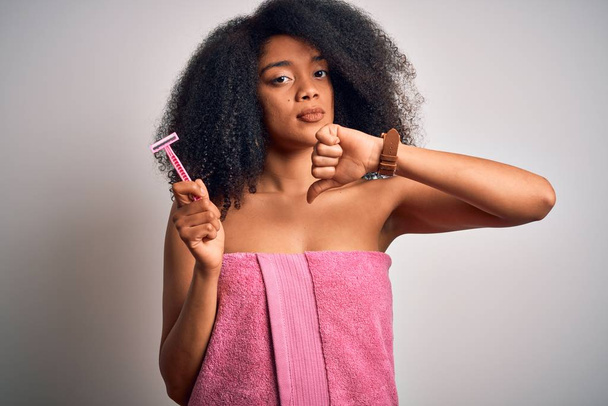 Giovane donna afro-americana con capelli afro in un asciugamano che tiene rasoio femminile per la depilazione del corpo con viso arrabbiato, segno negativo mostrando antipatia con i pollici verso il basso, concetto di rifiuto
 - Foto, immagini