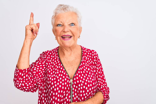 Femme âgée aux cheveux gris portant une veste décontractée rouge debout sur un fond blanc isolé pointant le doigt vers le haut avec une idée réussie. Sorti et heureux. Numéro un
. - Photo, image