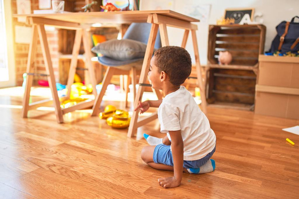 Hermoso niño afroamericano sentado en el suelo y sonriendo alrededor de un montón de juguetes en el jardín de infantes
 - Foto, Imagen