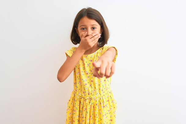 Jeune belle enfant fille portant une robe florale jaune debout sur fond blanc isolé se moquant de vous, pointant du doigt la caméra avec la main sur la bouche, expression de honte
 - Photo, image