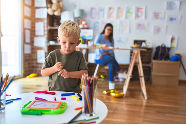 Молодой кавказский ребенок играет в детской школе с учителем. Мать и сын в игровой комнате рисуют ничью с цветными карандашами, молодая женщина на заднем плане сидит на столе
. - Фото, изображение