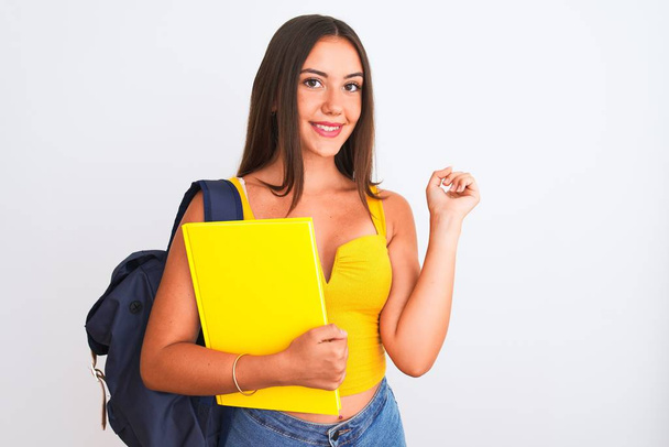 schöne Studentin trägt Rucksack hält Notizbuch über isolierten weißen Hintergrund sehr glücklich zeigt mit Hand und Finger zur Seite - Foto, Bild
