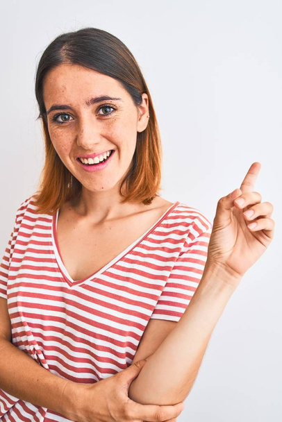 Hermosa mujer pelirroja vistiendo casual rayas camiseta roja sobre fondo aislado muy feliz señalando con la mano y el dedo a un lado
 - Foto, Imagen