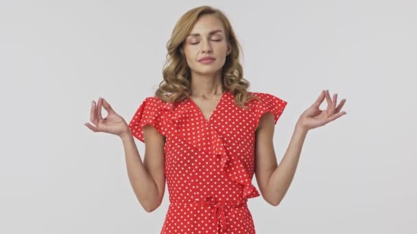 Carefree pretty blonde woman in dress meditating in yoga pose with closed eyes over grey background - Imágenes, Vídeo