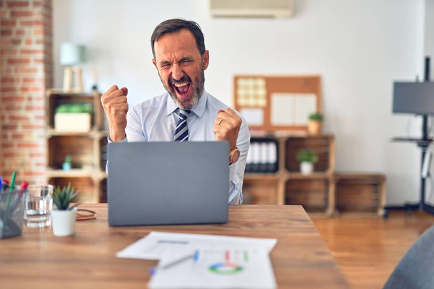 Középkorú jóképű üzletember nyakkendőt visel ül laptoppal az irodában nagyon boldog és izgatott csinál győztes gesztus felemelt karokkal, mosolyog és sikoltozik a sikerért. Ünnepi koncepció. - Fotó, kép