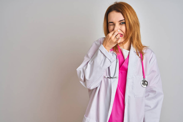 Redhead caucasian doctor woman wearing pink stethoscope over isolated background smelling something stinky and disgusting, intolerable smell, holding breath with fingers on nose. Bad smells concept. - Fotoğraf, Görsel